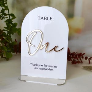 Könnte beinhalten: Ein bogenförmiges Tischschild aus weißem Acryl mit dem Wort TABLE in schwarzer Schrift, einer großen goldenen Schreibschrift One und dem schwarzen Text Thank you for sharing our special day. Das Schild steht auf einem transparenten Acrylsockel.