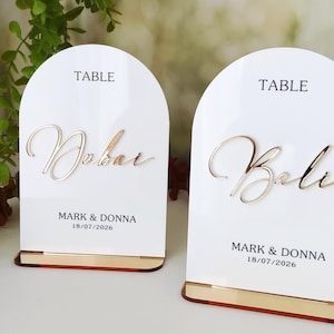 Puede incluir: Dos tarjetas de mesa de acrílico blanco con letras doradas. La palabra "TABLE" está impresa en la parte superior. Una tarjeta dice "Dubai" y la otra "Bali". Ambas tarjetas tienen los nombres "MARK & DONNA" y la fecha "18/07/2026" impresos debajo.