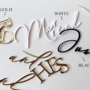 May include: Selection of wedding cake toppers in gold, white, and black. The toppers feature the words "Married", "Just", "Mr & Mrs", and "Hers & His". The words "Gold", "White", and "Black" are also visible.