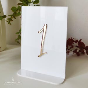 Puede incluir: Una tarjeta blanca con el número de mesa 1 en color dorado. La tarjeta es rectangular y se apoya sobre una base blanca. El número está en una fuente cursiva. Para decoración de bodas o eventos.