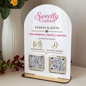 Könnte beinhalten: Ein weißes, bogenförmiges Acrylschild mit goldenen Akzenten, das den Text Sweetly Crafted, EVENTS & GIFTS und YOUR MOMENTS, SWEETLY CRAFTED zeigt. Es enthält goldene Instagram- und TikTok-Symbole, zwei quadratische QR-Codes und Social-Media-Namen.