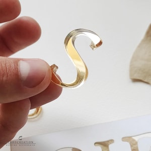 May include: A clear, gold-coloured letter "S" is held between fingers. The letter is made of a transparent material with a gold-coloured interior. The letter is part of a wedding accessory set. The brand name "CARDCREATION WEDDING ACCESSORIES" is visible.
