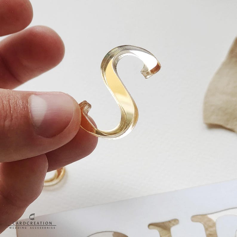 May include: A clear, gold-coloured letter "S" is held between fingers. The letter is made of a transparent material with a gold-coloured interior. The letter is part of a wedding accessory set. The brand name "CARDCREATION WEDDING ACCESSORIES" is visible.