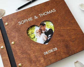 May include: A wooden wedding photo album with a heart-shaped cutout revealing a couple's photo. The album is inscribed with "SOPHIE & THOMAS" and the date "08/08/28". A black pen rests on the album's surface.