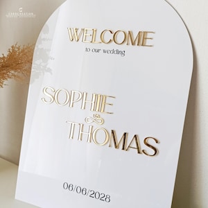 May include: White wedding sign with gold lettering. The sign says "WELCOME to our wedding" above the names "SOPHIE & THOMAS" and the date "06/06/2028". The sign has an arched top.