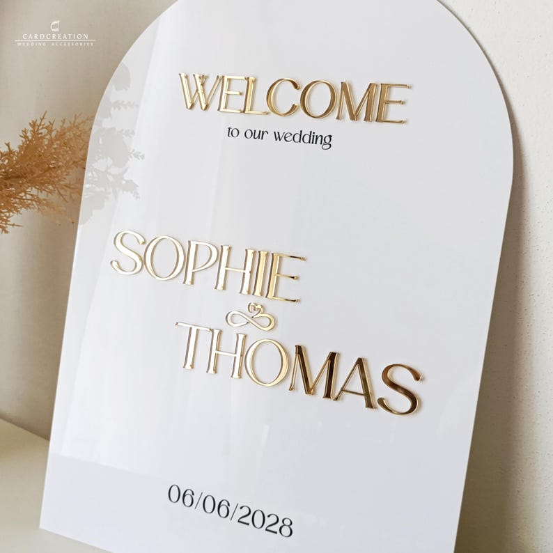 May include: White wedding sign with gold lettering. The sign says "WELCOME to our wedding" above the names "SOPHIE & THOMAS" and the date "06/06/2028". The sign has an arched top.
