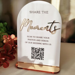 Peut inclure: Un panneau en acrylique blanc en forme d'arche avec une base dorée. Le texte indique SHARE THE MOMENTS, SCAN TO SHARE YOUR PHOTOS AND VIDEOS OF OUR WEDDING WITH US, et inclut un QR code noir et blanc. Le mot Moments est en lettres dorées.