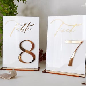 Può includere: Due segnaposto bianchi con dettagli dorati. Uno presenta la parola "Table" e il numero 8, l'altro la parola "Tisch" e il numero 7. Entrambi hanno una base dorata e sono progettati per matrimoni o eventi.