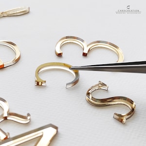 May include: Close-up of gold-coloured acrylic letters and numbers, including the number 3 and the letter S. A pair of tweezers is holding the letter C. The letters and numbers are for wedding accessories.
