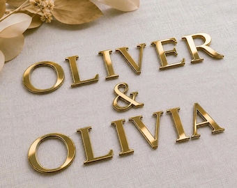 May include: Shiny gold-coloured acrylic letters spelling out OLIVER & OLIVIA in a serif font. The letters are arranged in three lines with the names separated by an ampersand symbol, featuring a reflective metallic finish on a light fabric.