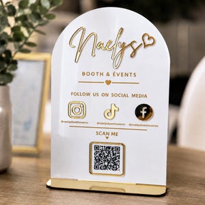May include: A white arched acrylic sign with gold lettering and icons. It features the text Naelys Booth & Events, social media icons for Instagram, TikTok, and Facebook with handles, and a central QR code above the text Scan Me on a gold base.