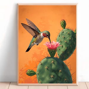 Könnte beinhalten: Gerahmter Kunstdruck mit einem Kolibri, der sich von einer rosa Blüte auf einem grünen Kaktus ernährt. Der Hintergrund ist orange. Der Kolibri hat grüne, rote und weiße Federn. Der Kaktus hat scharfe Stacheln.