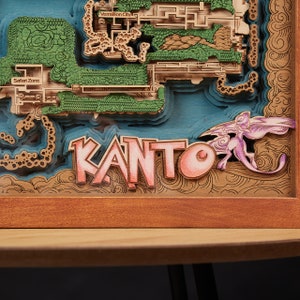 May include: A layered wooden artwork depicting a map of the Kanto region, with the word "KANTO" in pink letters. The map includes the Safari Zone and Vermillion City, with green and blue accents. A purple creature is on the right.