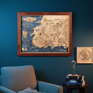 May include: A framed wooden map with a detailed, laser-cut design. The map features a landmass with intricate details, including rivers, mountains, and coastlines, set against a blue background. The frame is a rich, dark brown. A smaller, similar artwork is also visible.