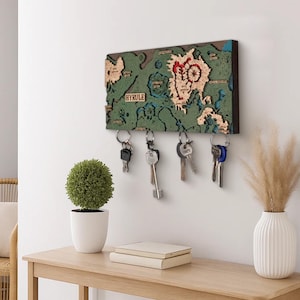 Può includere: Un portachiavi da parete rettangolare in legno con una mappa topografica a strati di Hyrule nei toni del verde, beige e rosso. La mappa include il testo "HYRULE" e quattro ganci in metallo, ognuno dei quali sostiene un anello portachiavi argentato.