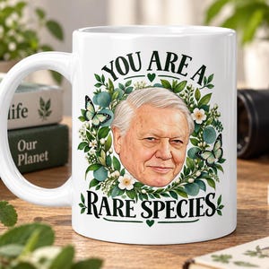 May include: A white ceramic mug featuring a portrait of David Attenborough surrounded by a wreath of green leaves, white flowers, and butterflies. The text "YOU ARE A RARE SPECIES" is printed in black lettering above and below the portrait.