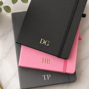 May include: Three personalised notebooks stacked. The top notebook is black with gold initials "DG", the middle one is pink with gold initials "HB", and the bottom one is grey with silver initials "TP". Each notebook has an elastic band.