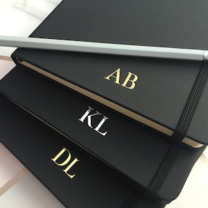 May include: Three black notebooks stacked on top of each other. The top notebook has the initials "AB" in gold lettering. The middle notebook has the initials "KL" in silver lettering. The bottom notebook has the initials "DL" in gold lettering.