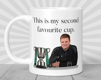 Peut inclure: Une tasse en céramique blanche avec une image en noir et blanc d'un homme tenant un trophée. La tasse porte l'inscription "This is my second favourite cup."