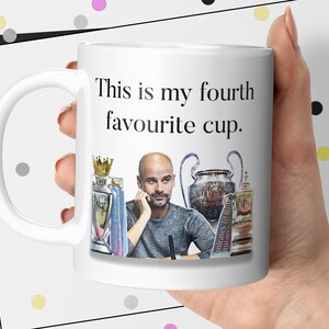 K&ouml;nnte beinhalten: Wei&szlig;e Keramik-Tasse mit dem Text "This is my fourth favourite cup." und einem Bild eines Mannes und Troph&auml;en. Die Tasse hat einen gebogenen Henkel und wird von einer Person gehalten.