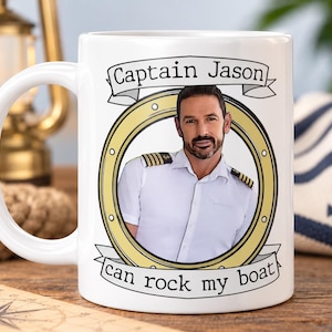 May include: A white ceramic mug featuring a circular graphic of a person in a captain's uniform inside a gold-coloured porthole frame. Above the frame is a banner with the text "Captain Jason" and below is a banner with "can rock my boat".