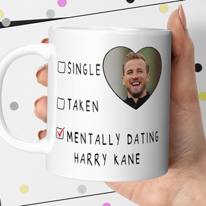 Pu&ograve; includere: Tazza in ceramica bianca con la scritta "Single", "Taken" e "Mentally Dating Harry Kane". Una foto di Harry Kane &egrave; all'interno di una forma a cuore. La tazza &egrave; tenuta da una persona.
