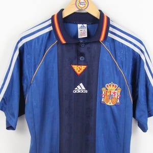 May include: Blue and navy short-sleeved soccer jersey with white stripes on the sleeves. Features the Adidas logo, a crest, and a small emblem. The jersey is hanging on a wooden hanger.