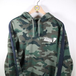 Könnte beinhalten: Grüner Camouflage-Hoodie mit schwarzen Seitenteilen und einem weißen Patch mit dem Text "TRUE ORIGINALS" und "EST. 1998".