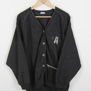 May include: A dark gray cardigan with a zippered pocket and button closure. The cardigan has a V-neck and the text "DRIFT" is embroidered on the front.