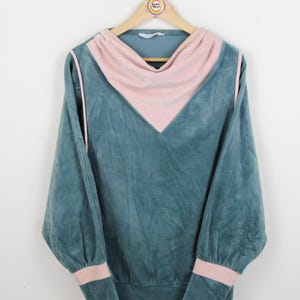 Puede incluir: Una sudadera de terciopelo azul turquesa con cuello y ribetes de terciopelo rosa. La sudadera tiene mangas largas y un corte holgado.