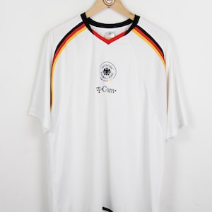 Op de afbeelding: Wit voetbalshirt met zwarte, rode en gele accenten. Het heeft een V-hals en het logo van de Duitse voetbalbond met "T-Com" eronder gedrukt. Het shirt hangt aan een houten hanger.