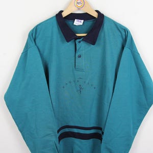 Pode incluir: Camisa polo de manga comprida verde-azulado com gola e punhos azul marinho. A camisa apresenta o texto "EAGLE CLUB" em uma fonte azul escuro na frente. Duas listras azul marinho estão na parte inferior da camisa.