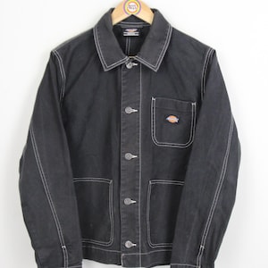 May include: A black denim work jacket with white stitching and a small orange and brown logo patch on the left chest. The jacket has a button-up front and multiple pockets.