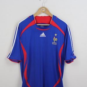 May include: A blue football shirt with red and white detailing, displaying the Adidas logo and the French Football Federation emblem. The shirt has short sleeves and is suspended from a wooden hanger.