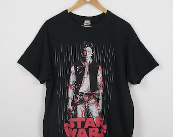 Puede incluir: Camiseta negra con un gráfico de Han Solo de Star Wars. El gráfico es en rojo, blanco y gris, y muestra a Han Solo de pie bajo la lluvia. El texto "STAR WARS" está impreso debajo del gráfico.