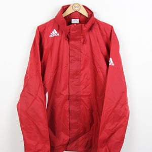 May include: A red Adidas jacket with a hood, hanging on a hanger. The Adidas logo is visible on the chest and sleeve. The jacket is made of a lightweight material.