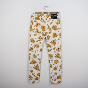 Puede incluir: Vaqueros blancos con un estampado barroco dorado. Los vaqueros cuelgan de una percha de madera. Se ve la etiqueta Versace Jeans Couture.