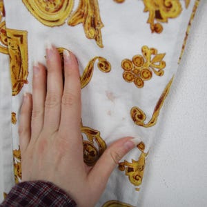 Puede incluir: Tejido vaquero blanco con un patr&oacute;n barroco dorado repetido. El tejido tiene manchas marrones y est&aacute; siendo tocado por una mano con u&ntilde;as largas. El tejido forma parte de una prenda.