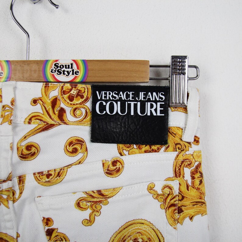 Puede incluir: Vaqueros blancos con un estampado barroco dorado. Los vaqueros tienen un parche de cuero negro con el texto "VERSACE JEANS COUTURE". Los vaqueros est&aacute;n colgados en una percha de madera con el texto "Soul & Style".