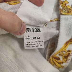 Puede incluir: Primer plano de tejido vaquero blanco con estampado barroco dorado. Se ve una etiqueta con "VERSACE JEANS COUTURE", junto con un c&oacute;digo de producto y una direcci&oacute;n web para comprobar la autenticidad.