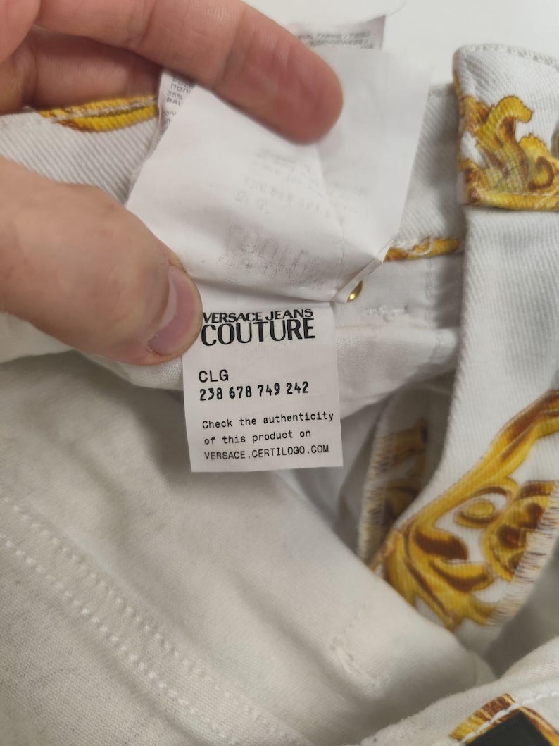 Puede incluir: Primer plano de tejido vaquero blanco con estampado barroco dorado. Se ve una etiqueta con "VERSACE JEANS COUTURE", junto con un c&oacute;digo de producto y una direcci&oacute;n web para comprobar la autenticidad.