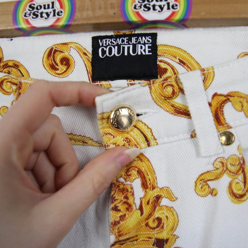 Puede incluir: Primer plano de unos vaqueros blancos con un estampado barroco dorado. Los vaqueros tienen botones dorados y una etiqueta negra que dice "VERSACE JEANS COUTURE". Los vaqueros forman parte de una colecci&oacute;n de ropa.