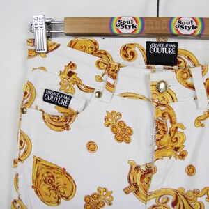 Puede incluir: Vaqueros blancos con un estampado barroco dorado. Los vaqueros presentan un dise&ntilde;o cl&aacute;sico de cinco bolsillos, cierre de bot&oacute;n y cremallera, y la etiqueta "Versace Jeans Couture". Los vaqueros cuelgan de una percha de madera con el texto "Soul & Style".