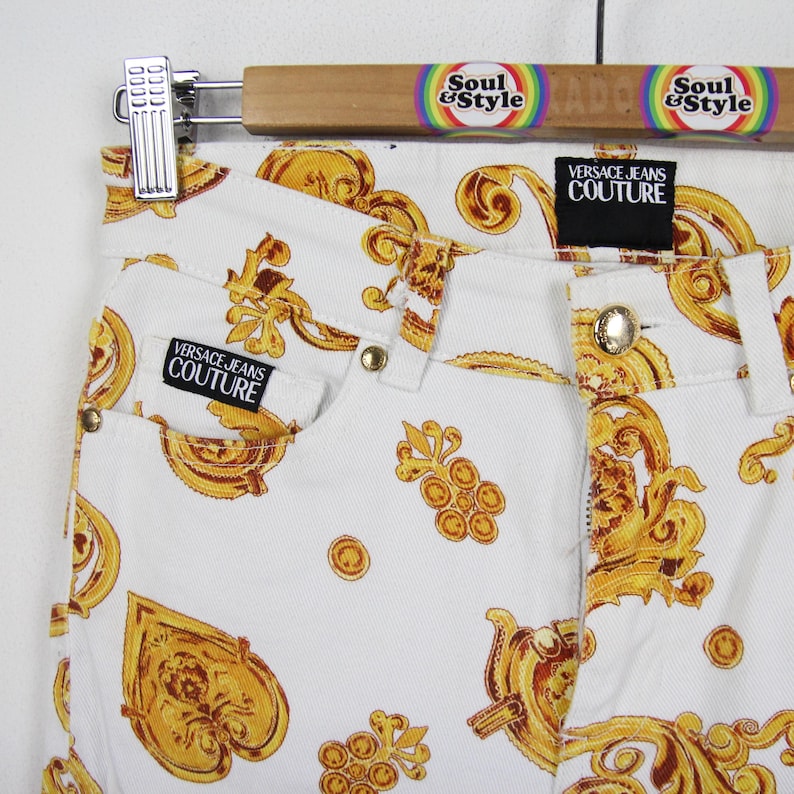 Puede incluir: Vaqueros blancos con un estampado barroco dorado. Los vaqueros presentan un dise&ntilde;o cl&aacute;sico de cinco bolsillos, cierre de bot&oacute;n y cremallera, y la etiqueta "Versace Jeans Couture". Los vaqueros cuelgan de una percha de madera con el texto "Soul & Style".