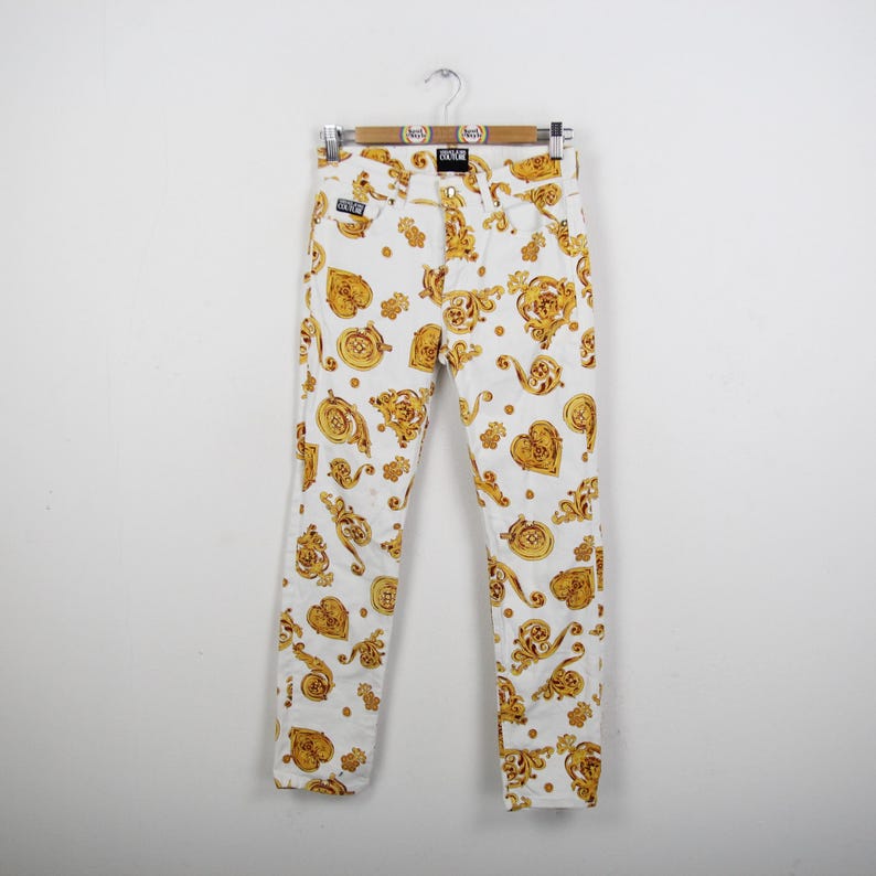 Puede incluir: Pantalones vaqueros blancos con un estampado barroco dorado. Los pantalones cuelgan de una percha de madera. El estampado dorado incluye corazones, medallones y otros elementos decorativos.