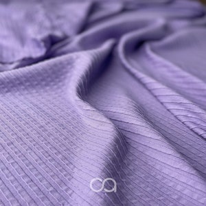 Könnte beinhalten: Nahaufnahme eines weichen, strukturierten, lilafarbenen Stoffes mit einem gerippten Muster. Der Stoff ist gefaltet und drapiert und zeigt seine Textur und Farbe.