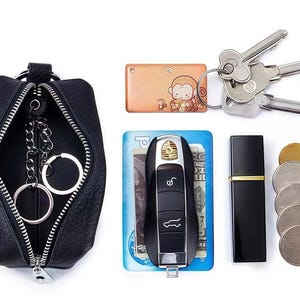 May include: A black leather key pouch with a silver zipper, containing key rings. Also shown are keys, a car key, a small perfume bottle, a coin purse, and a keychain with a monkey design.