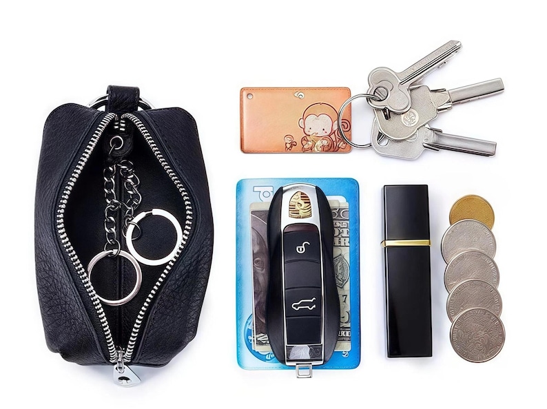 May include: A black leather key pouch with a silver zipper, containing key rings. Also shown are keys, a car key, a small perfume bottle, a coin purse, and a keychain with a monkey design.