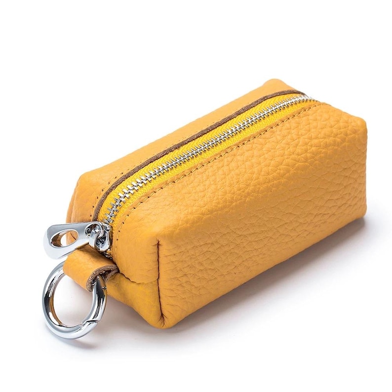 May include: Mustard yellow leather key case with a silver zipper and key ring. The rectangular case has a textured surface and a brown accent along the zipper. The zipper is yellow and the key ring is silver.