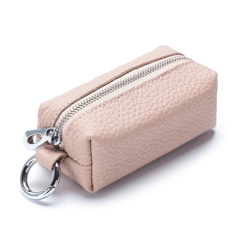 May include: A small, rectangular, blush-colored leather key pouch with a silver zipper and a silver key ring. The pouch has a textured surface and a light-colored interior. The zipper pull is also silver.